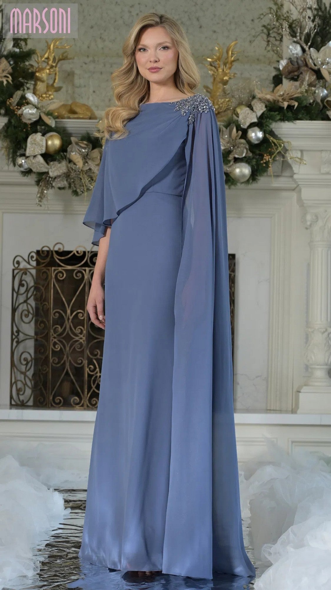 A woman in the Marsoni MV1362 long formal dress in slate blue with cape sleeves and crystal-beaded embellishment stands before a white fireplace decorated with greenery and ornaments.